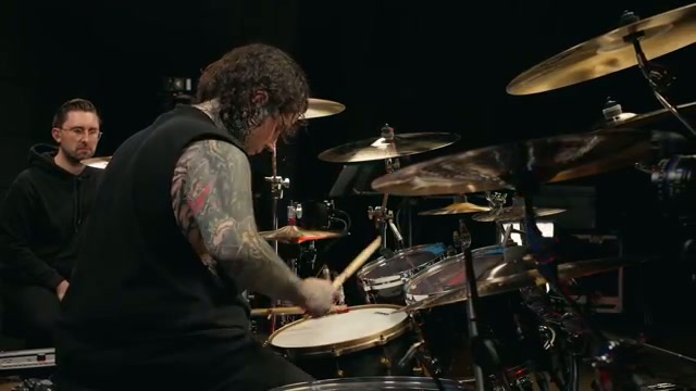 Austin Archey toca la batería visto desde atrás mientras el presentador de Drumeo reacciona al fondo