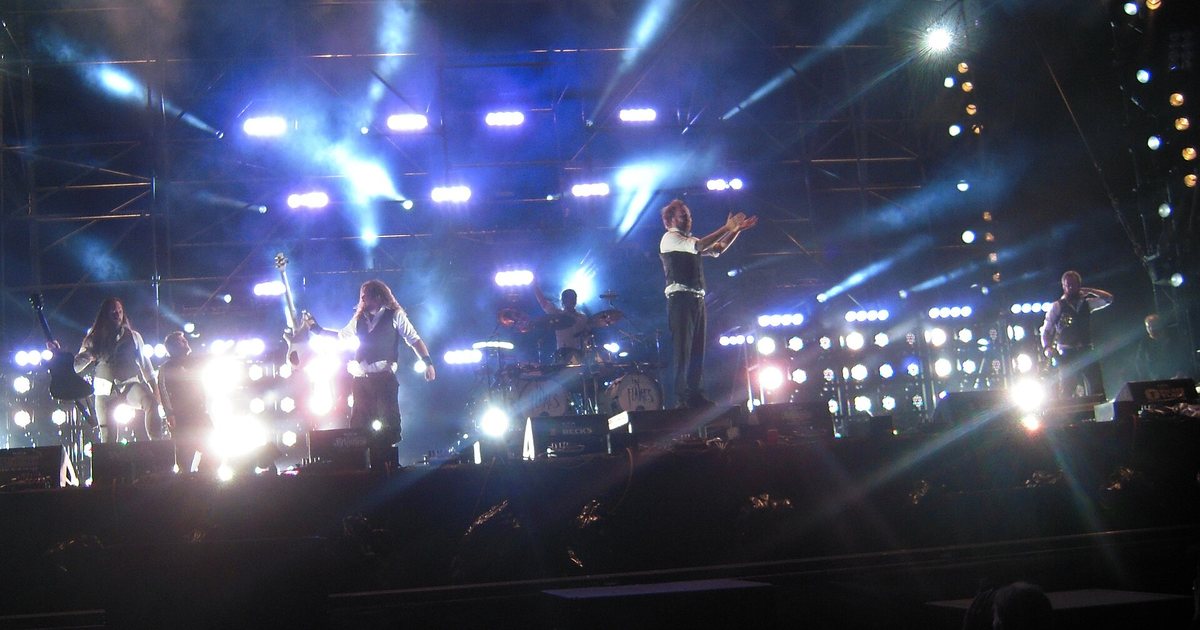 In Flames en un escenario de festival visto de frente, con el nombre de la banda en el bombo y potentes luces azules y blancas